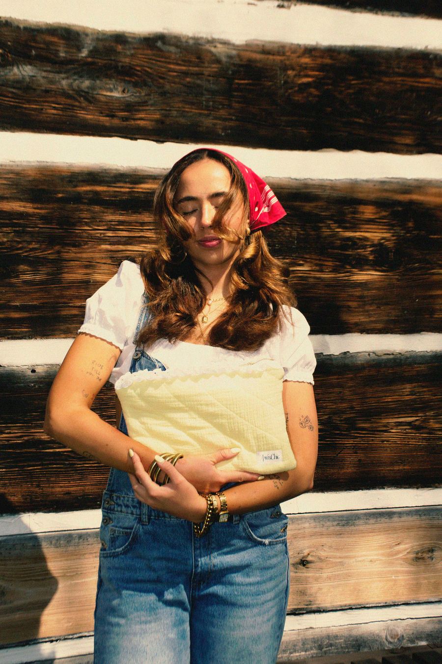 Model with eyes closed in denim overalls and white top, red bandana in her hair, holding a pale yellow quilted clutch against a wooden log wall.