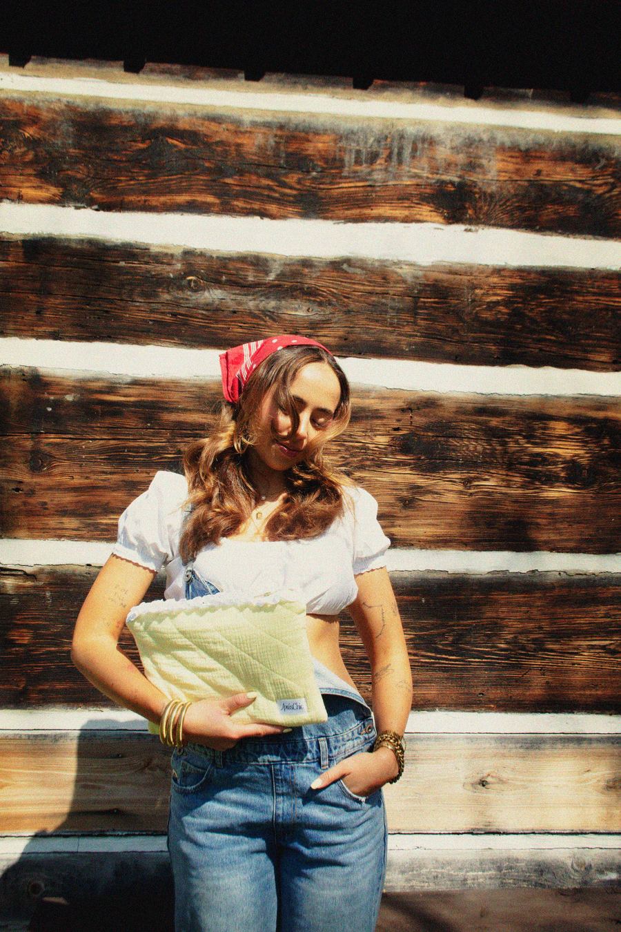 Model looking down and smiling softly, in white top and denim overalls with a red bandana, hand in her jeans pocket, holding a yellow quilted clutch.