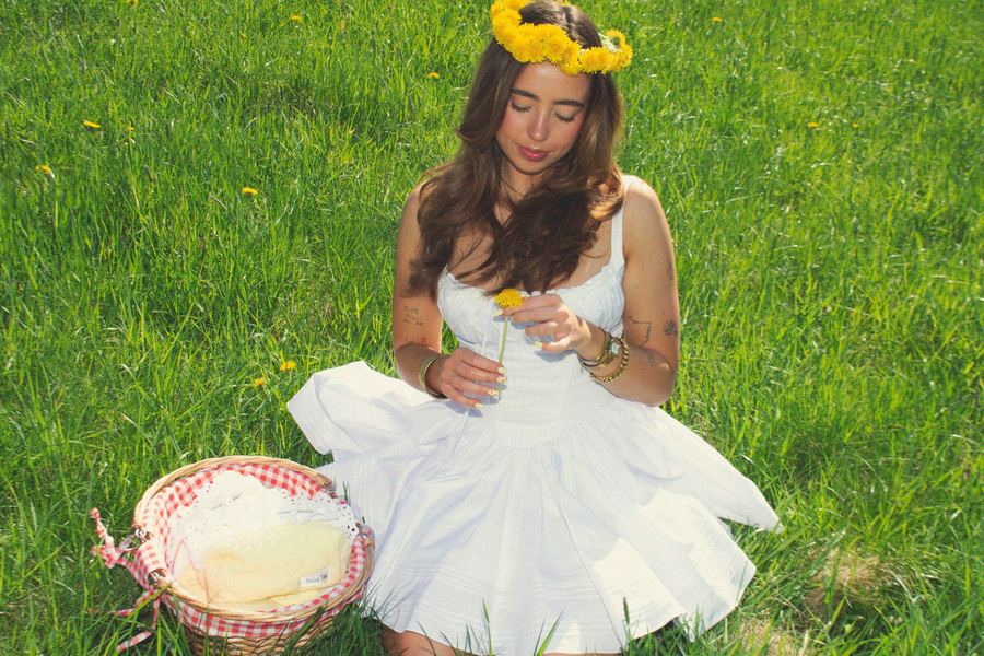 Seated model in a white sundress with a dandelion flower crown, holding a dandelion in the grass beside a red-gingham-lined picnic basket.