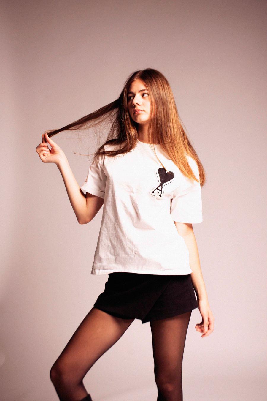 Model in an oversized white graphic tee, black skort, and sheer tights, mid-motion pulling a hand through her hair.