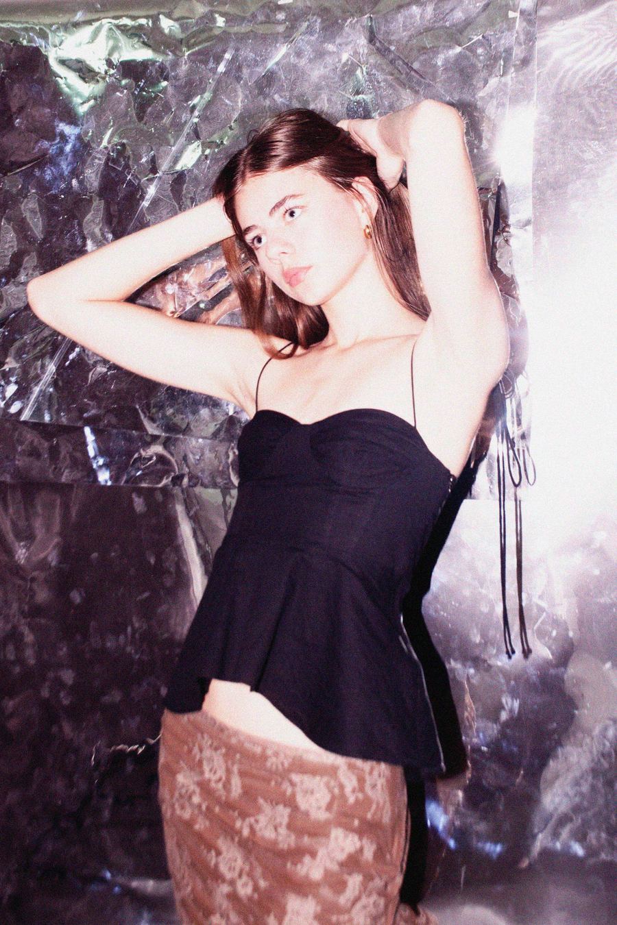 Model in a black bustier top and shimmering skirt, arms raised behind her head, against a crumpled foil backdrop.