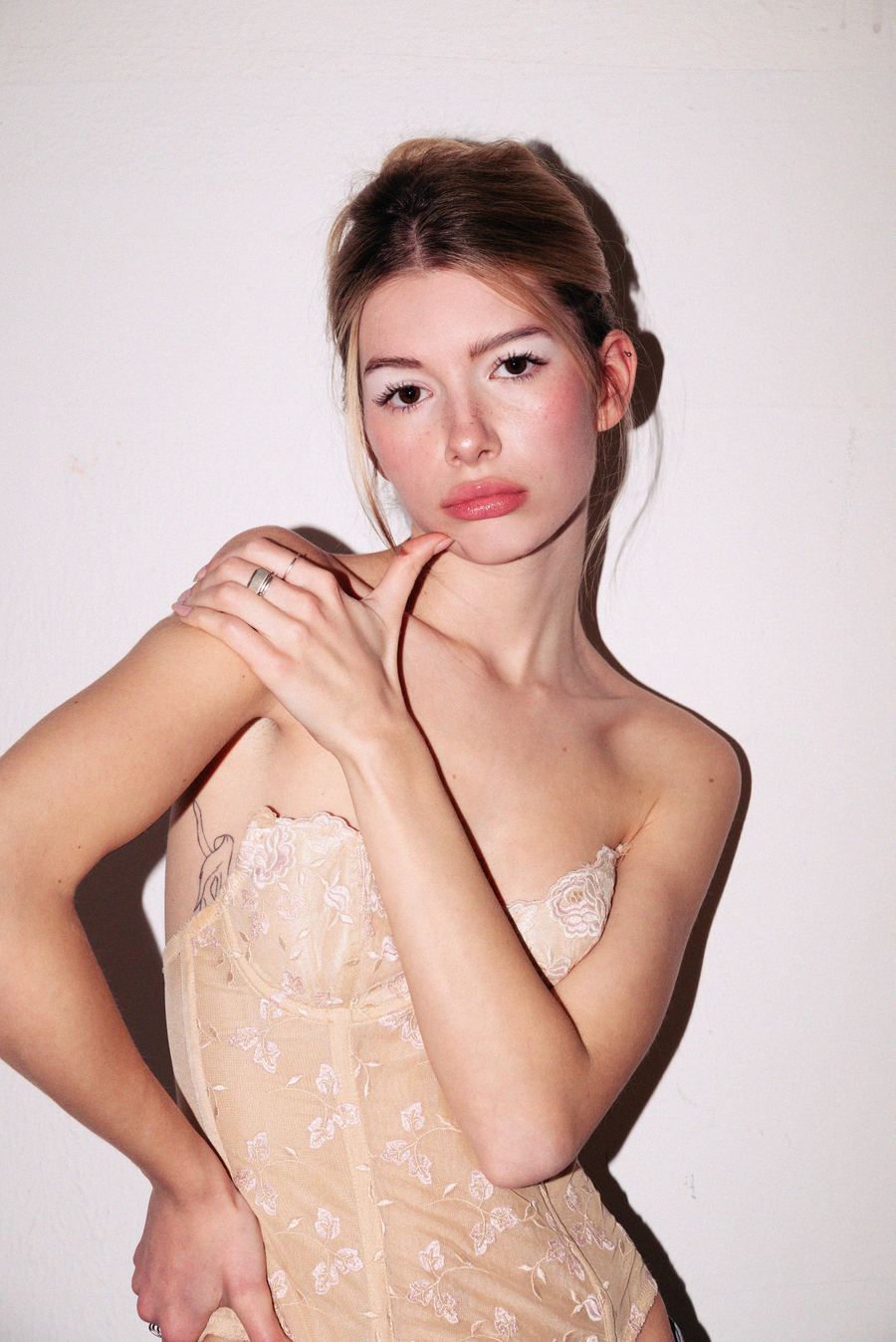 Strapless cream lace corset top, hand raised to the collarbone, small rib tattoo, plain white wall.