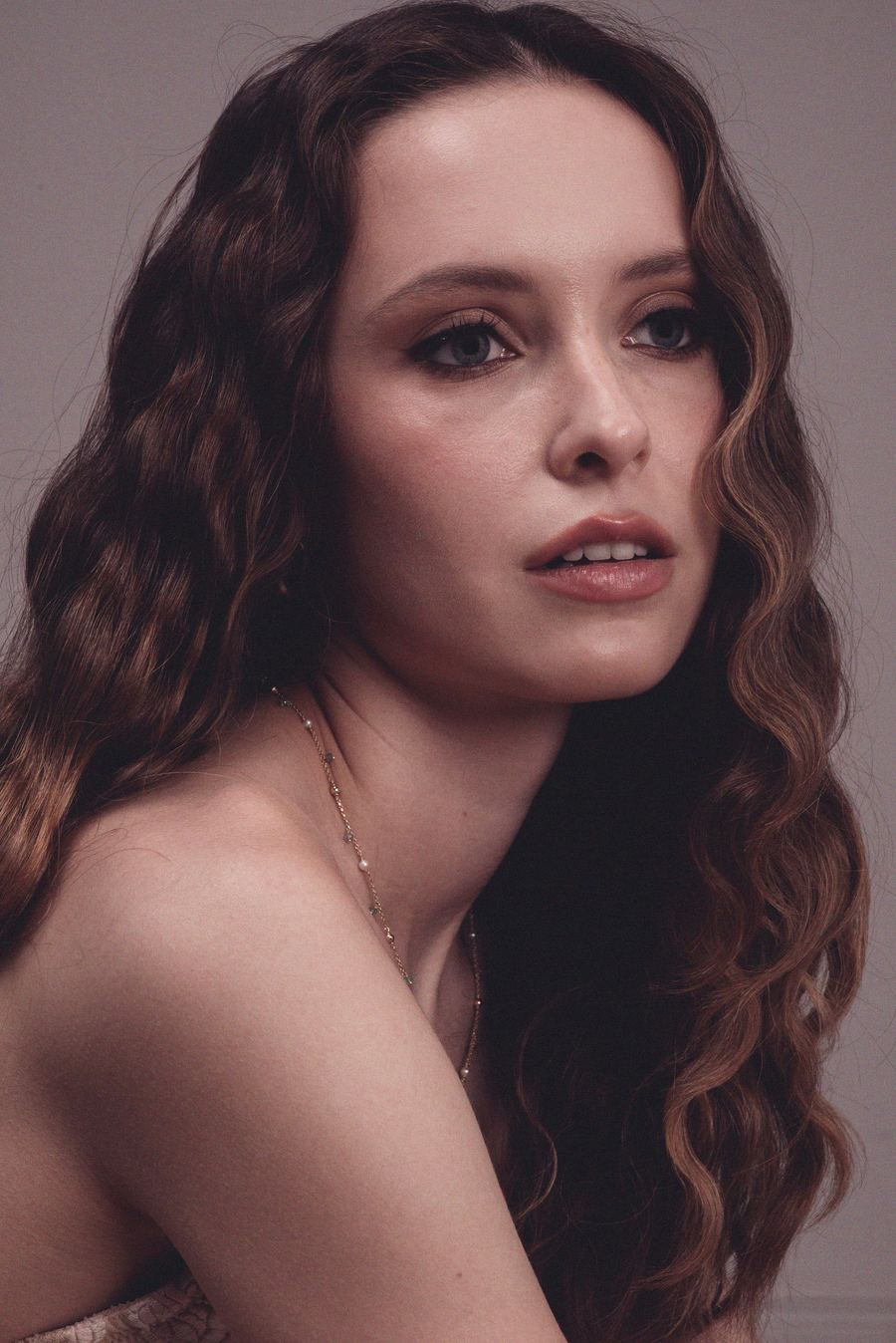 Close portrait, dark curly hair, delicate beaded necklace, bare shoulder, parted lips, warm light.