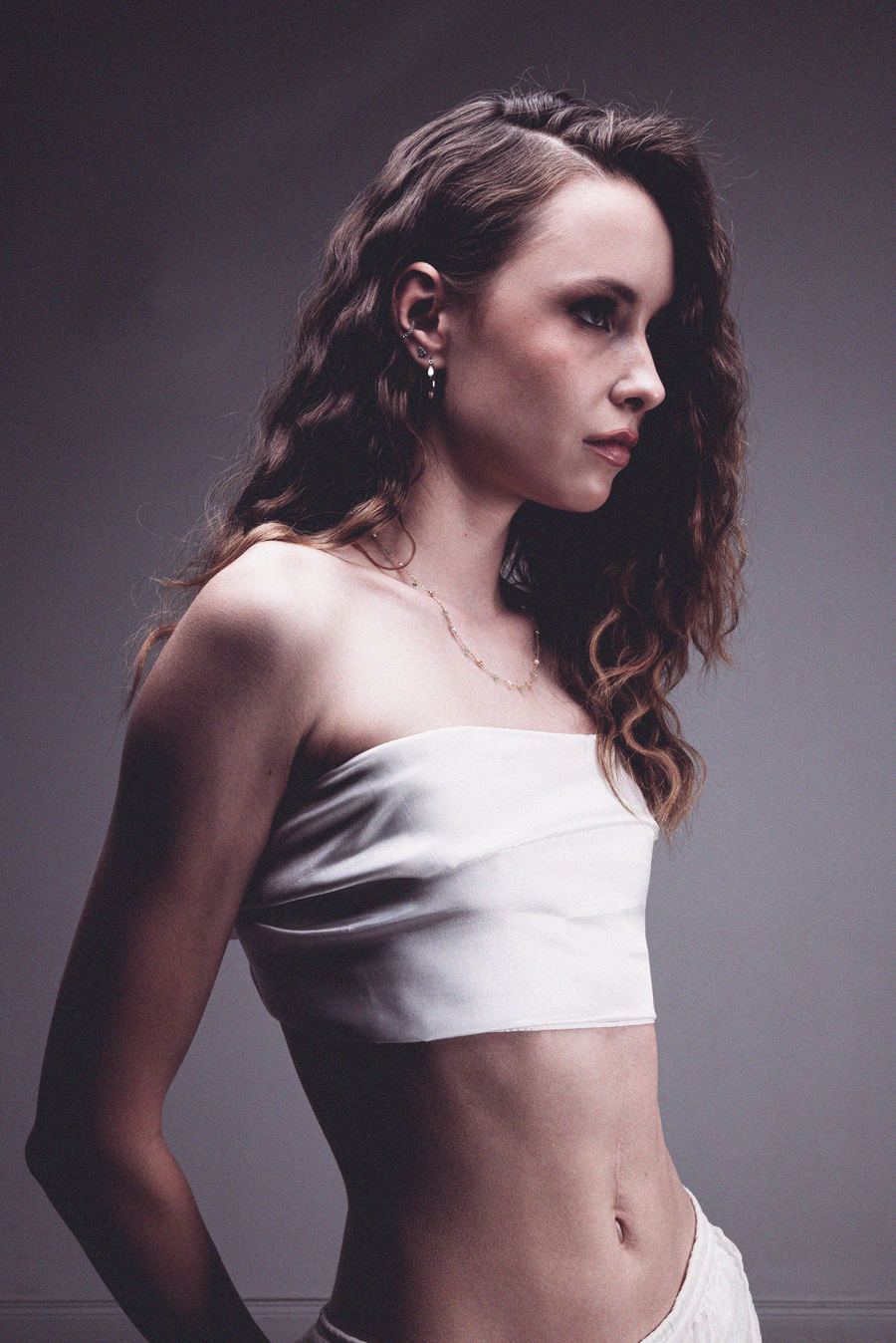 Profile portrait, curly hair, white strapless top, layered necklaces, drop earrings, gray backdrop.