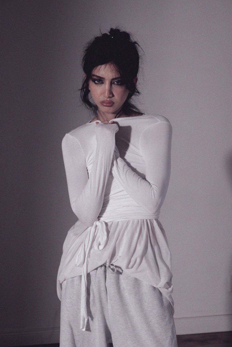 Portrait in a white wrap top, sleeves pulled over the hands and held near the chin, gray wall.