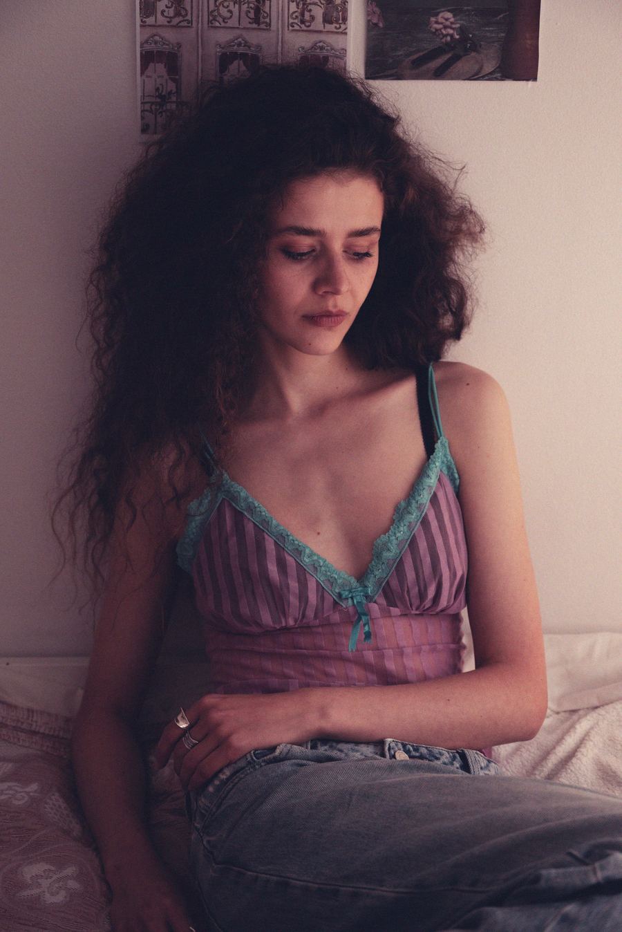 Seated on a bed in a purple striped cami with teal lace trim, looking down, posters on the wall.