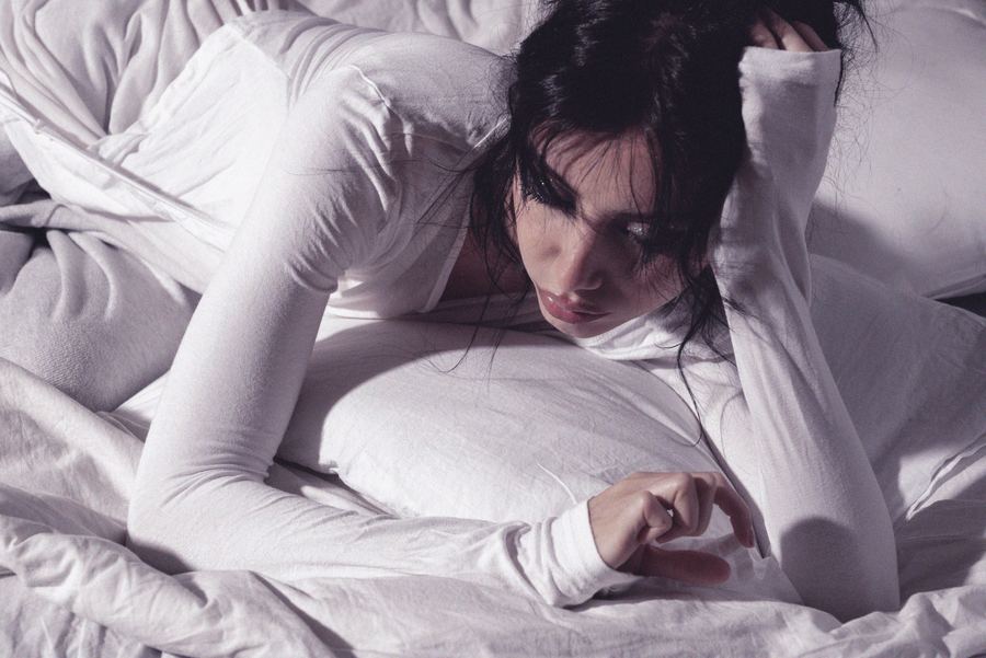 Lying in white bedding in a white long-sleeve top, hand raised, gazing downward, low light.
