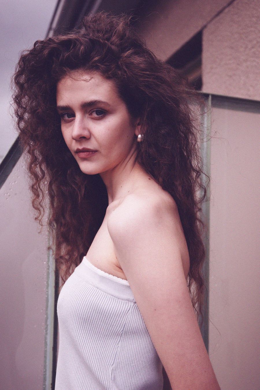 Profile portrait against a pale wall with a glass panel, long curly hair, white ribbed top.