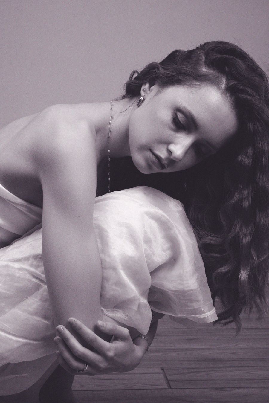 Black-and-white image, curled forward with knees drawn up, flowing white top, eyes closed, wood floor.