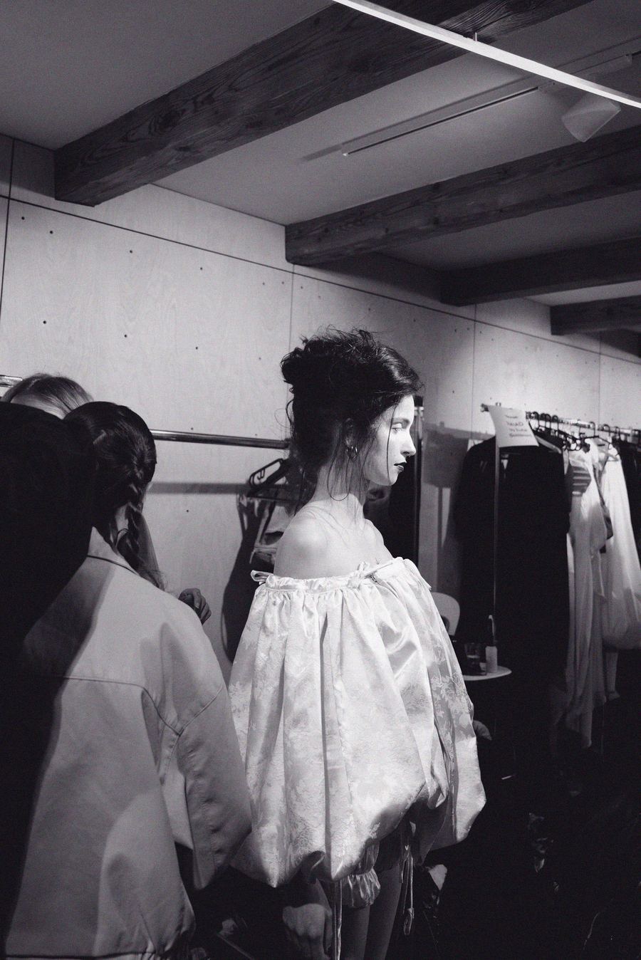 Backstage, a dresser adjusts a model's off-the-shoulder satin dress with a voluminous pleated skirt.