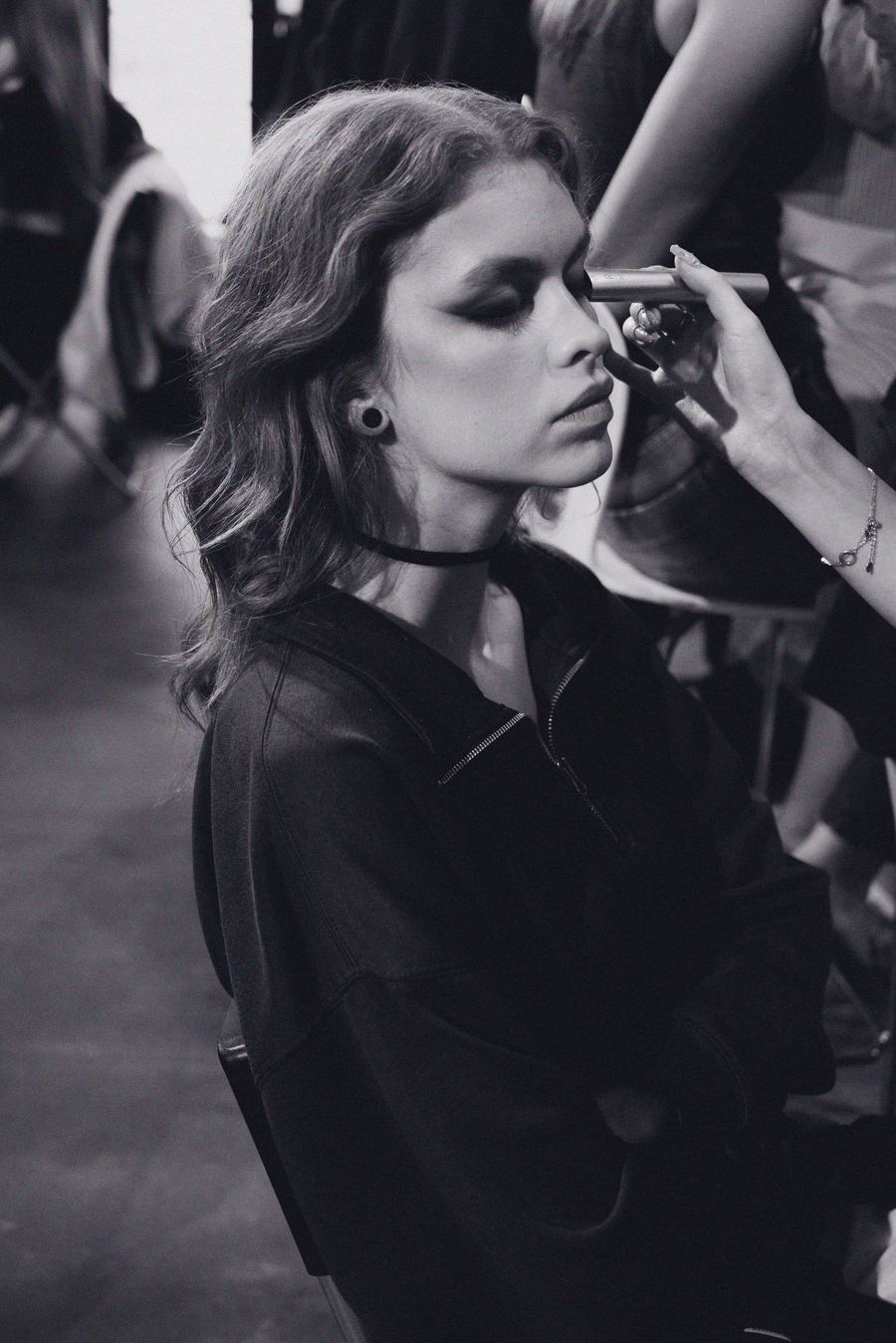 Backstage, a makeup artist applies eyeshadow to a model with a black choker and dark wavy hair.