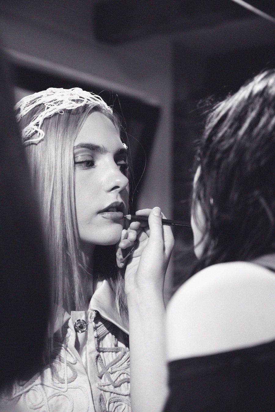 Backstage, a stylist lines a model's lips; she wears a woven thread headpiece and an embroidered coat.