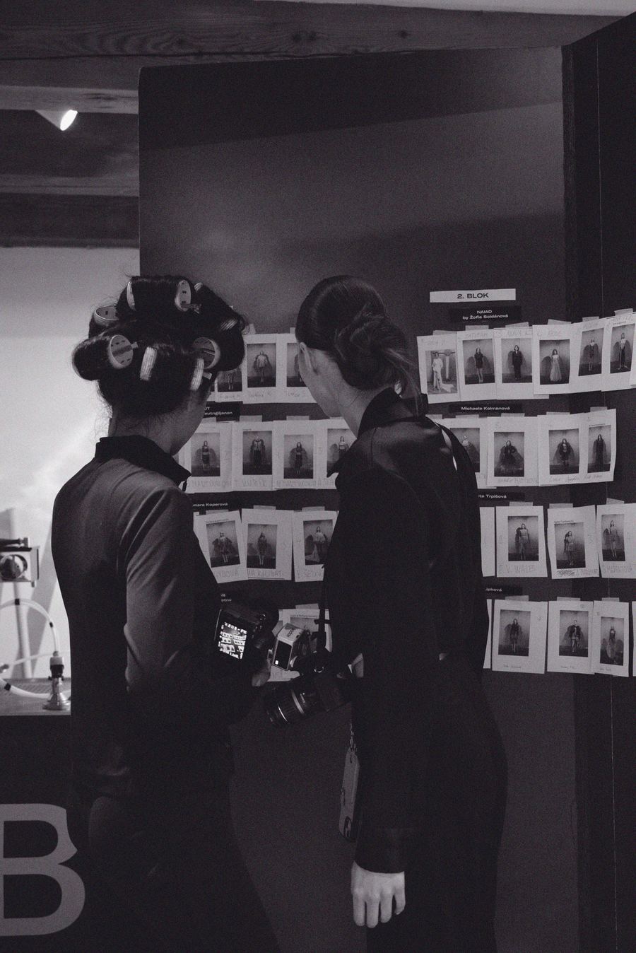 Backstage, two crew members with cameras review a wall of model casting polaroids before the show.