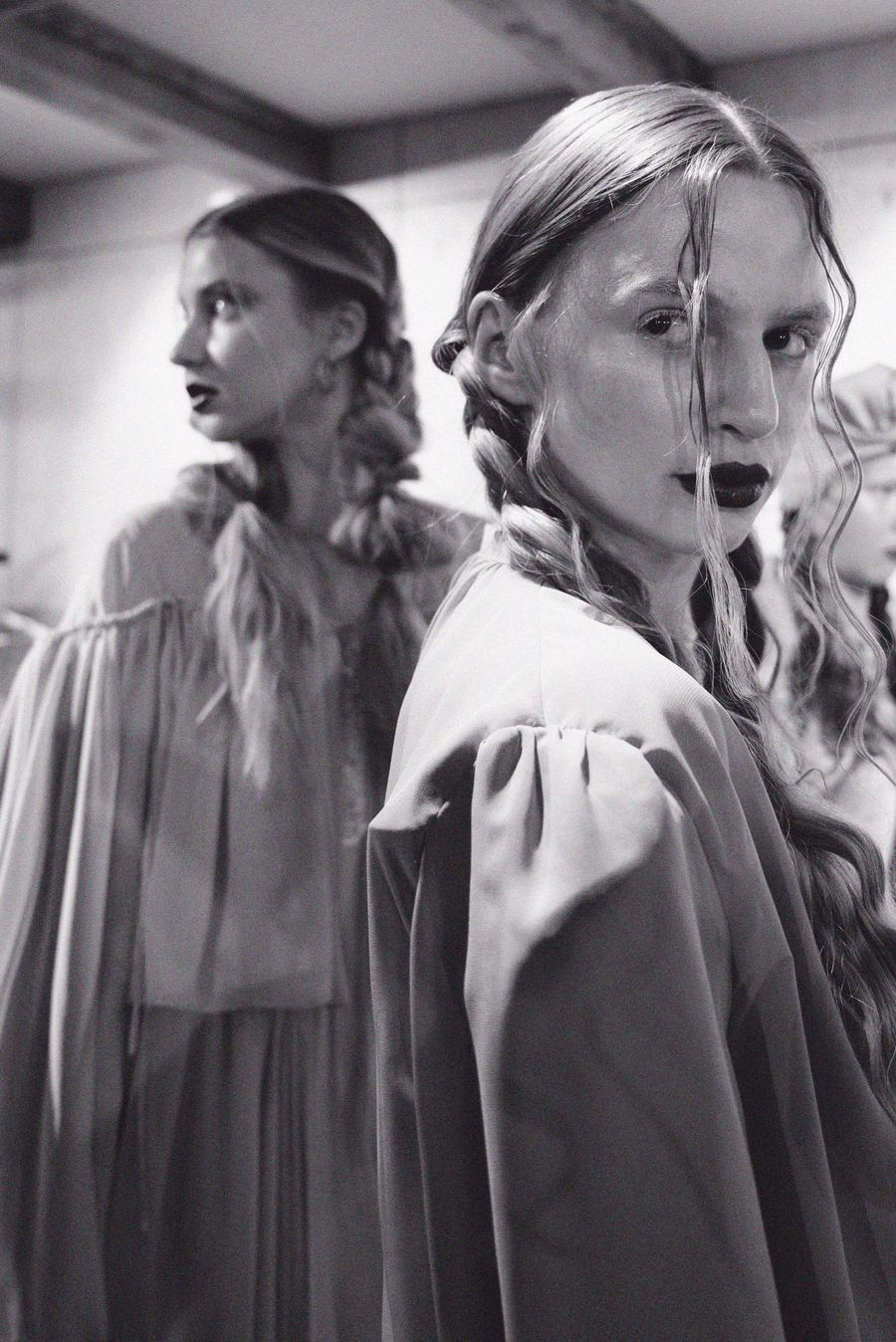 Two models in puff-sleeve satin tops stand back to back backstage, one gazing directly at the camera.