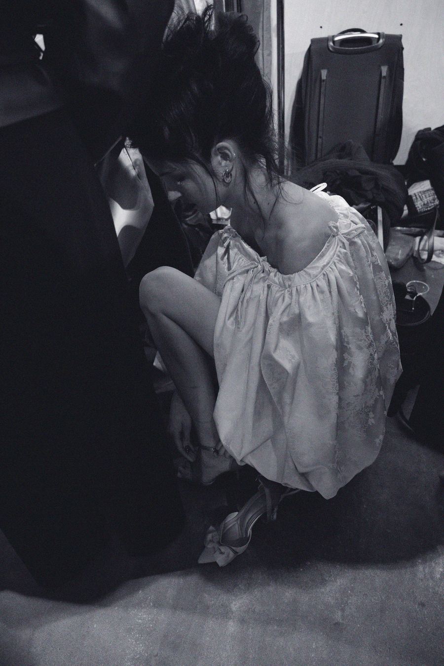 Backstage, a model crouches in a cream lace balloon-hem dress and bow-detail heels near a suitcase.