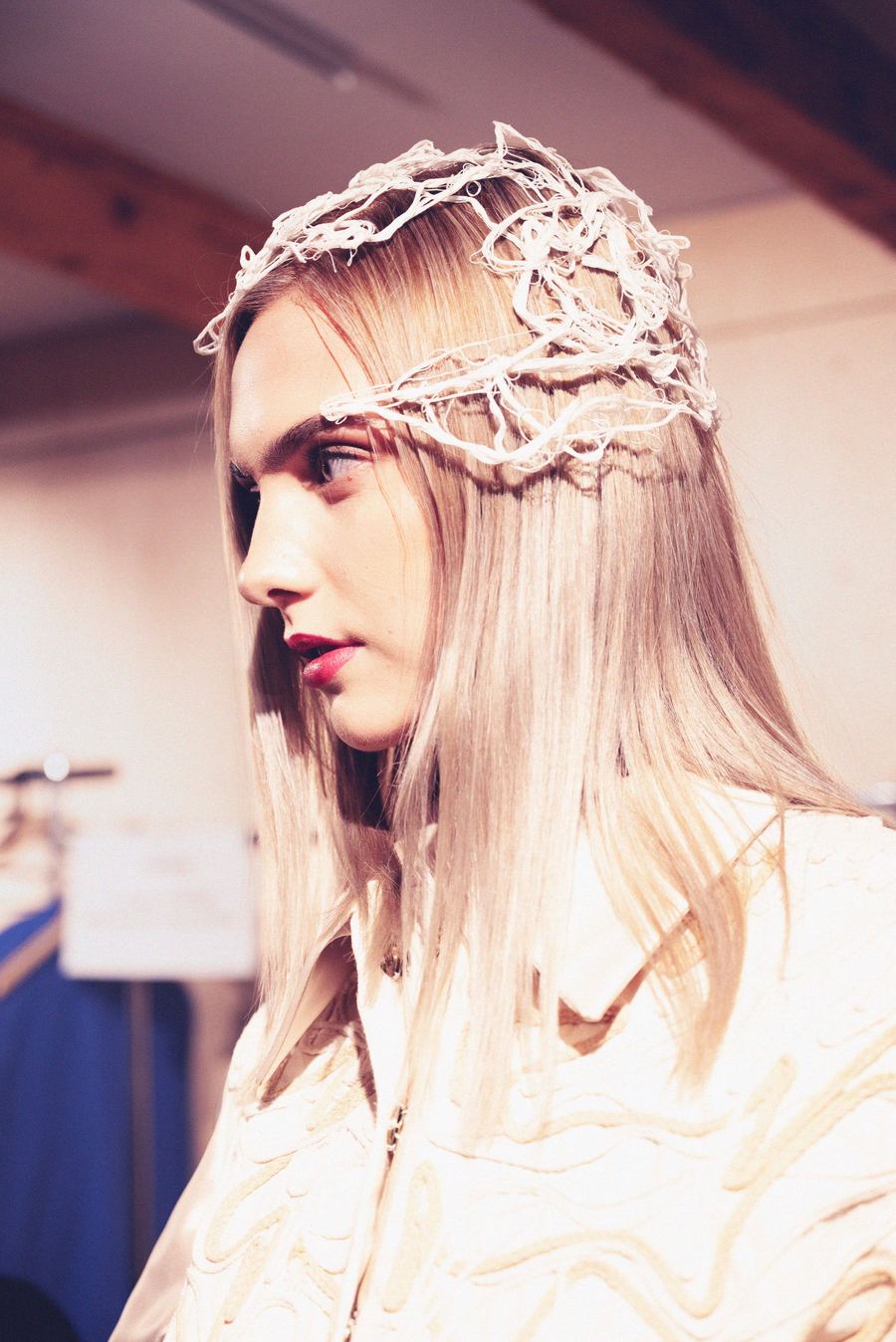 A model in profile wears a woven thread headpiece and dark red lipstick, backstage before the show.