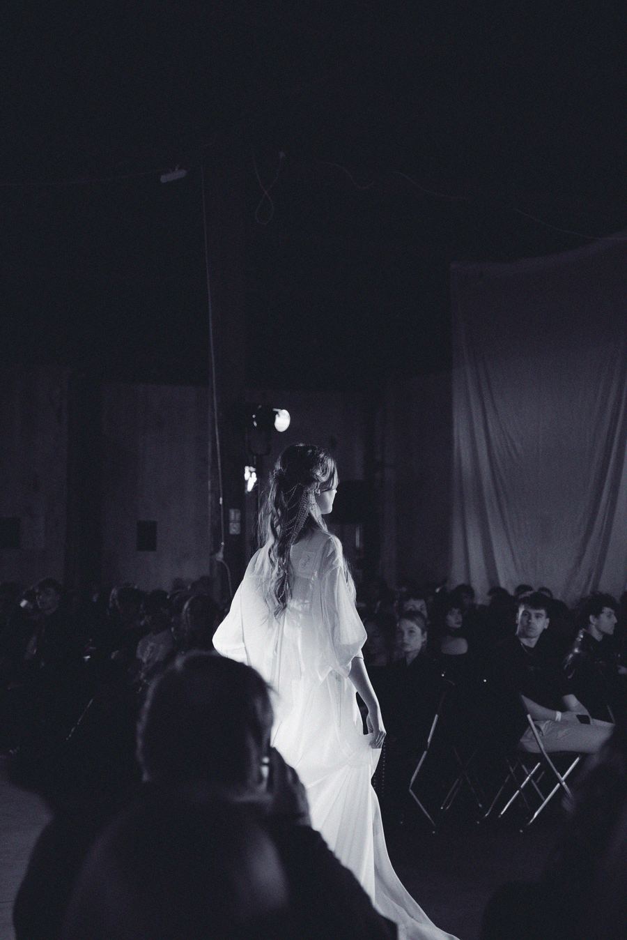 A model in a flowing white gown with wide sleeves walks the runway, audience seated in the dim light.