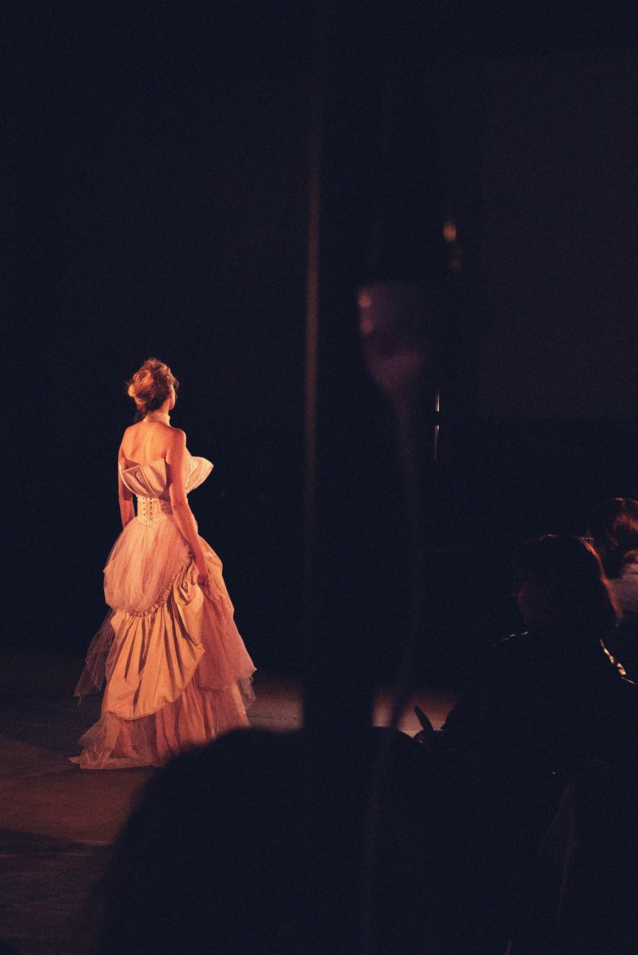 On the runway, a model in a blush tulle gown with a corseted back and tiered ruffled skirt walks away.
