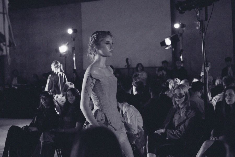 A model in a structured off-shoulder bodice walks the runway past photographers and seated guests.