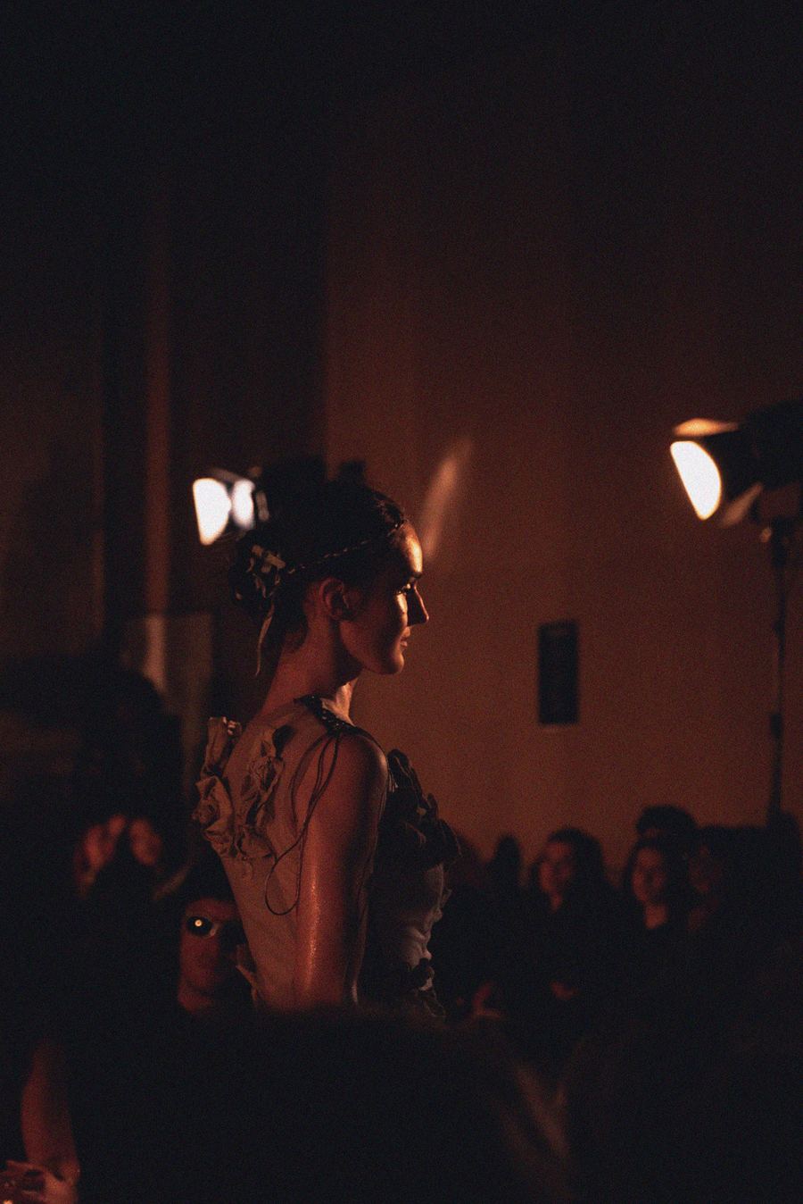 On the runway, a model in a floral lace dress with tie-back straps walks past the audience in warm light.