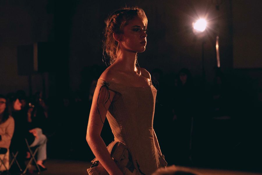 A model in a cream corseted off-shoulder gown walks the runway under bright stage lighting.