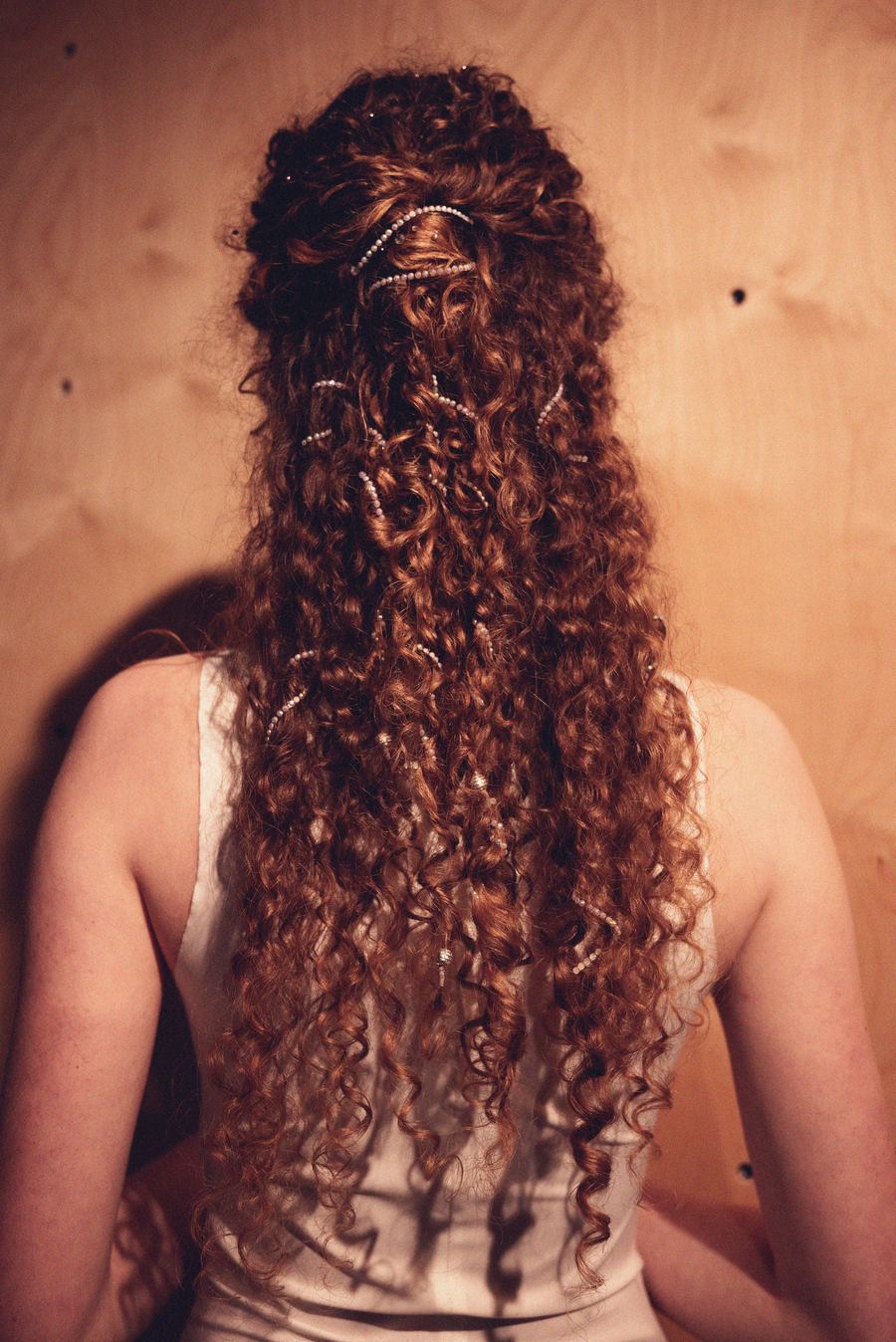 Model seen from behind, long curly hair threaded with small pearl strands, wearing a white sleeveless top backstage.