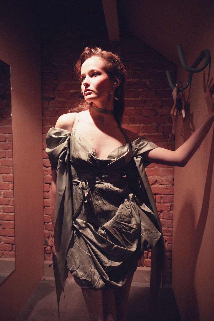Model in an off-shoulder olive taffeta dress with a corseted, gathered skirt, standing against a brick wall backstage.