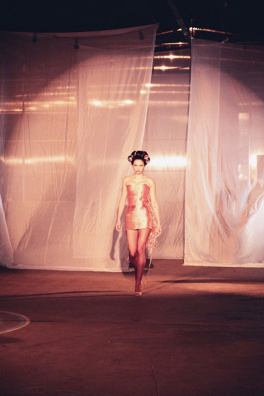 Model in curlers walks the runway in a strapless metallic bronze mini dress with fringed side detailing.