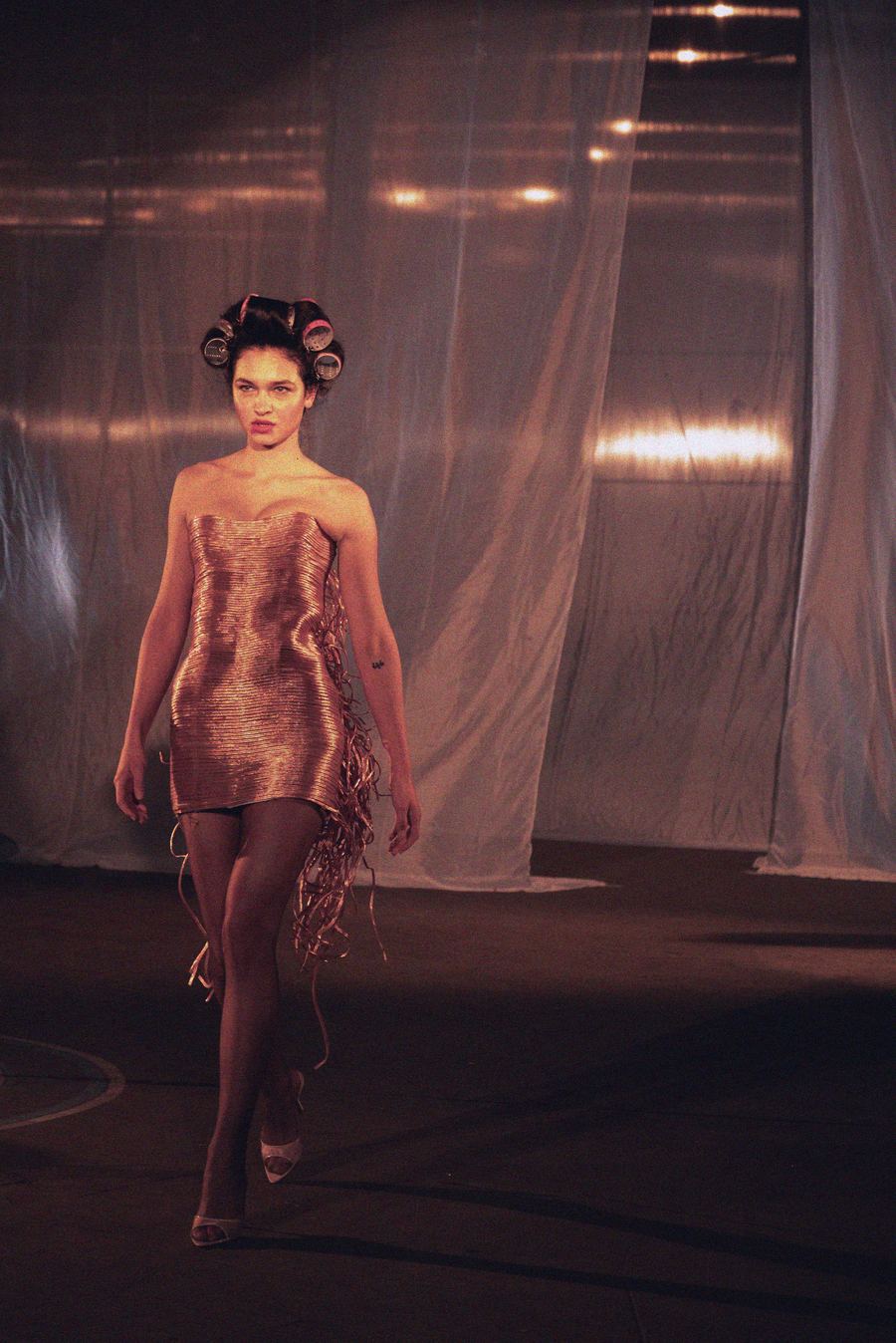 Model with curlers still in her hair walks the runway in a strapless pleated metallic gold mini dress with dangling fringe.