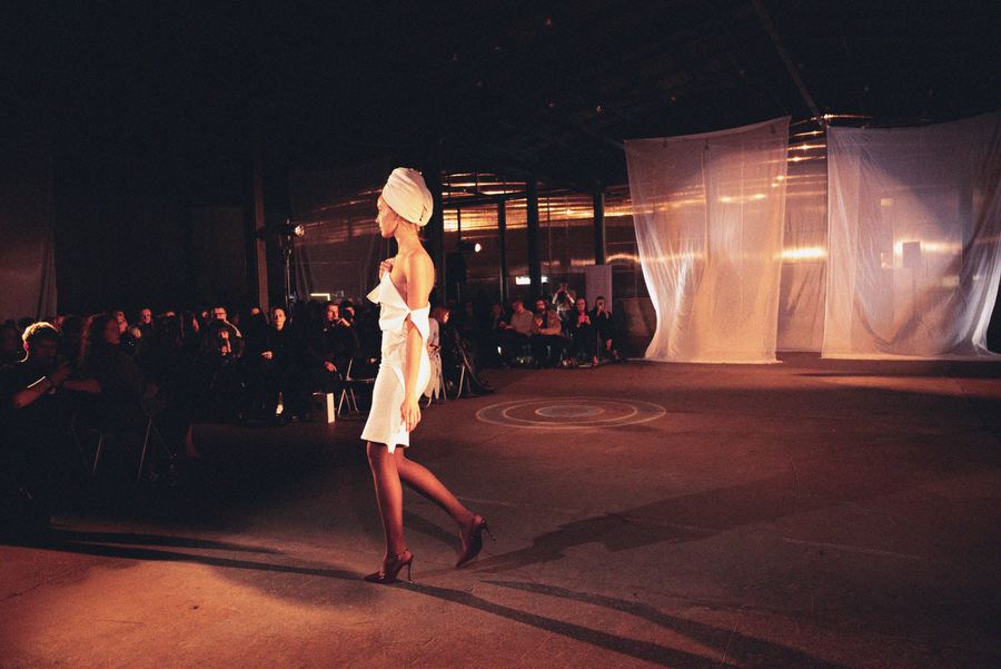 Model with a towel wrapped around her head walks the runway in a white draped strapless dress, seen from the side.