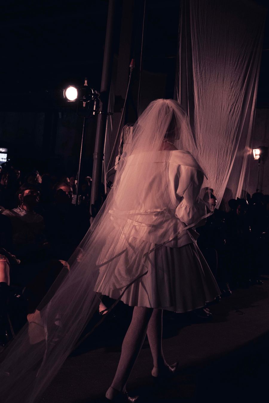 Model in a full tulle veil and puff-sleeved white dress walks away from the camera on the runway.