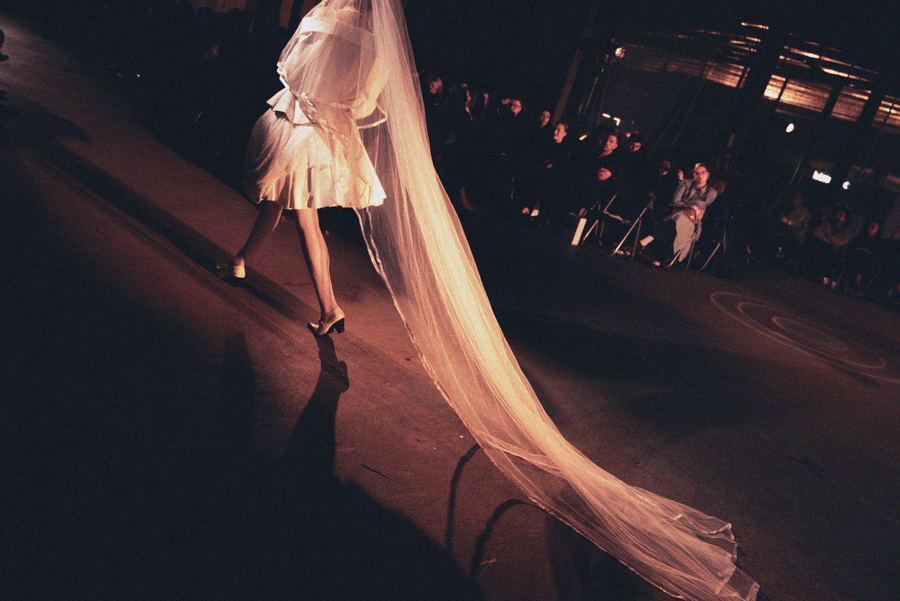Overhead view of a model in a short white pleated dress trailing a long sheer veil across the runway, audience seated at the edge.