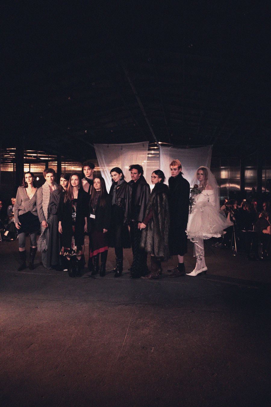 Finale lineup of models on the runway in dark tailored coats and layered black outfits, with a bride in white and veil at the end holding a rose.