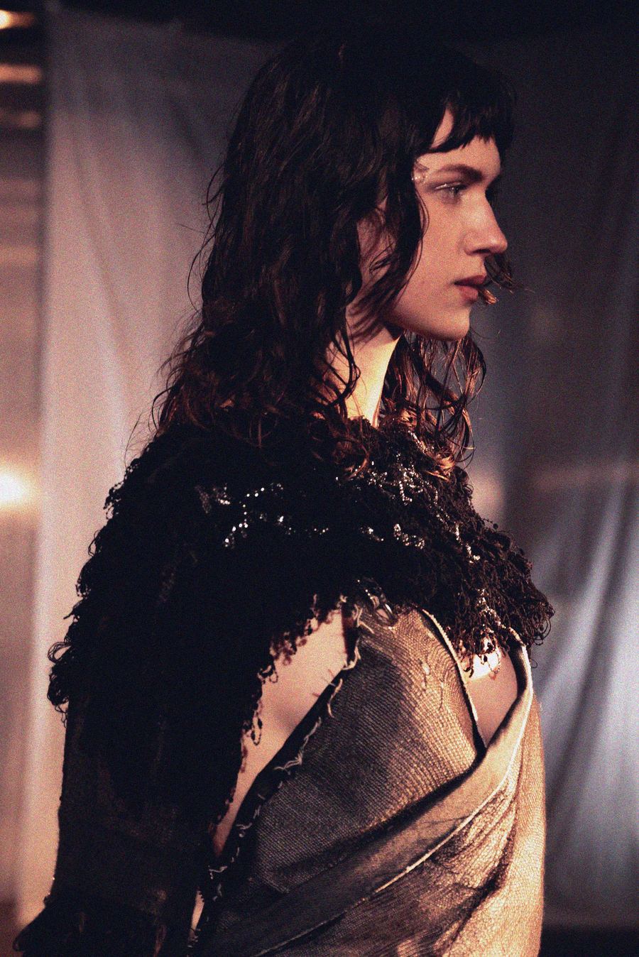 Close-up profile of a model with tousled dark hair wearing a black fringed, sequin-trimmed top over a draped metallic wrap.