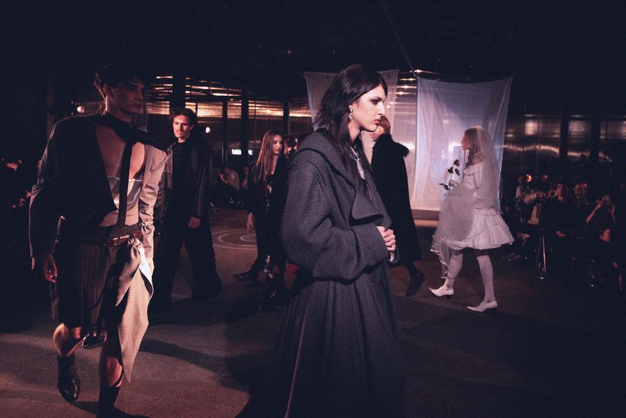 Models cross paths backstage, one in a dark hooded wool coat, another in a sheer cutout top, with a bride in white veil visible behind.