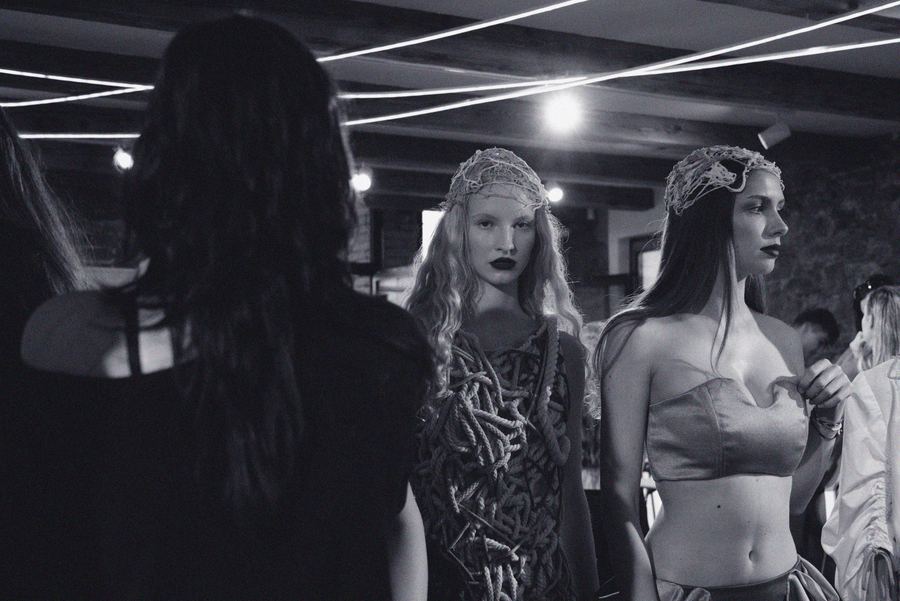 Two models backstage in netted, wire-textured headpieces, one in a rope-draped top and the other in a strapless satin bustier.