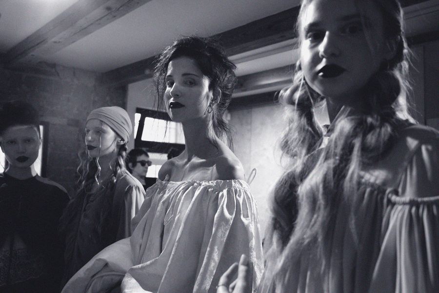 Group of models backstage with dark lipstick, one wearing a draped satin turban-style headpiece, others in scarves and off-shoulder gathered tops.