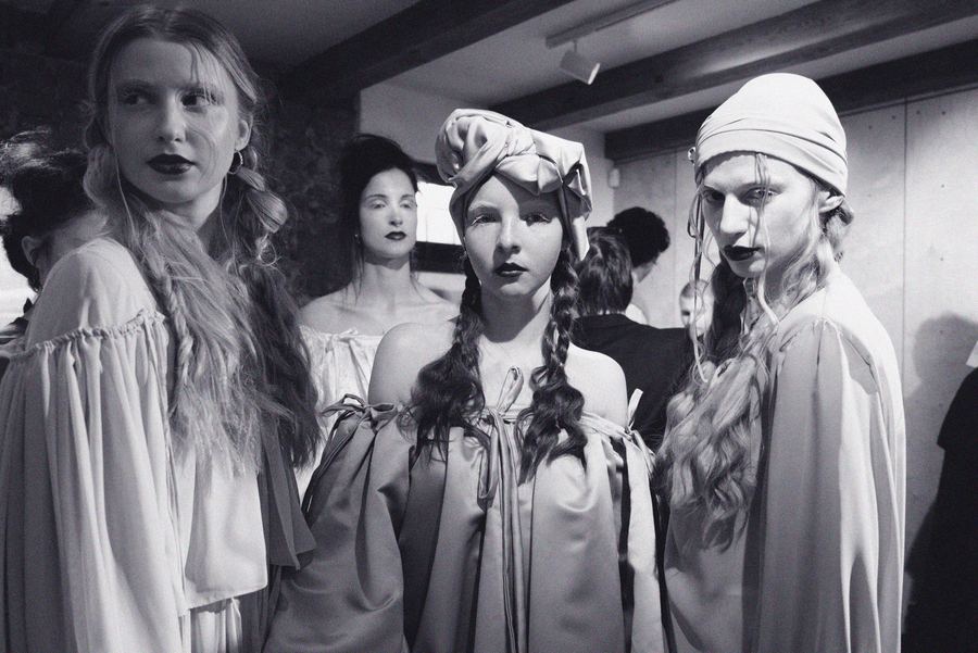 Three models backstage in pale draped off-shoulder gowns, headwrapped in fabric turbans, with dark lipstick and braided hair.