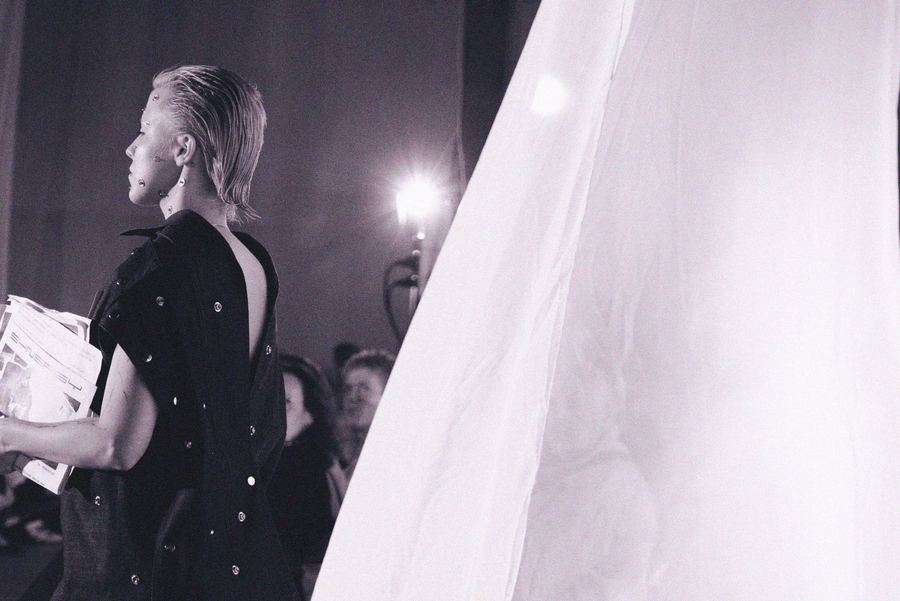 Profile of a model with slicked-back platinum hair and face piercings, wearing a black backless top with metal stud details, backstage beside a white drape.