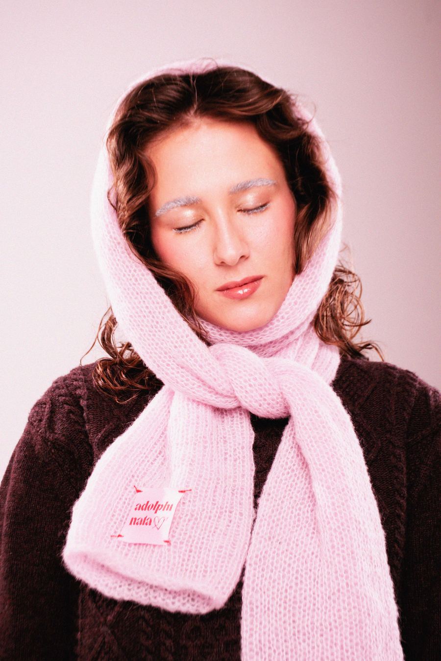 Model with eyes closed, wrapped in a chunky pink knit hood scarf over a dark brown cable-knit sweater.