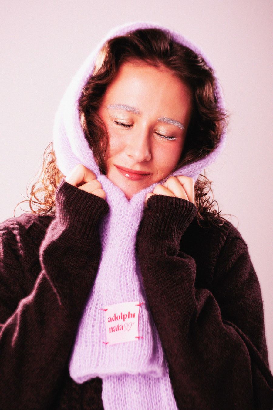 Model in a lilac knit hood scarf, hands tucked into an oversized brown coat, eyes closed and smiling softly.