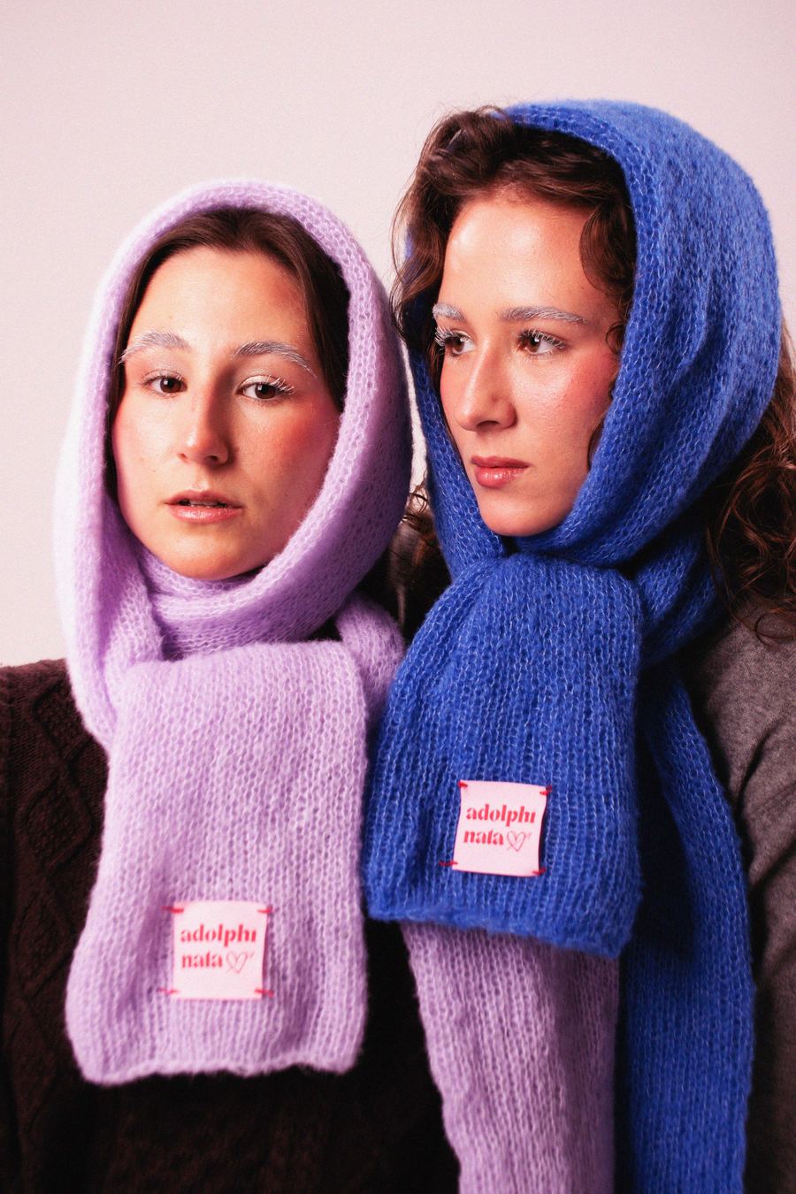 Two models side by side in knit hood scarves, one lilac and one cobalt blue, worn over sweaters.