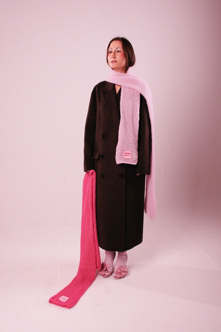 Model in a long brown double-breasted coat with a pink knit scarf, holding a hot-pink knit wrap, in pink heels.