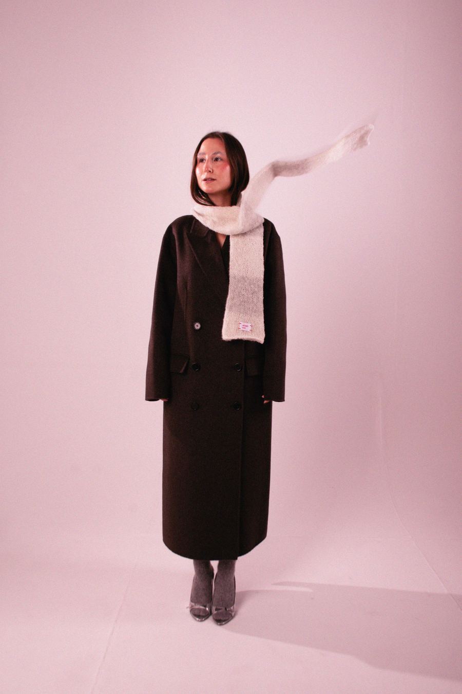 Model in a dark brown coat with a cream knit scarf caught mid-motion, paired with gray pointed-toe heels.