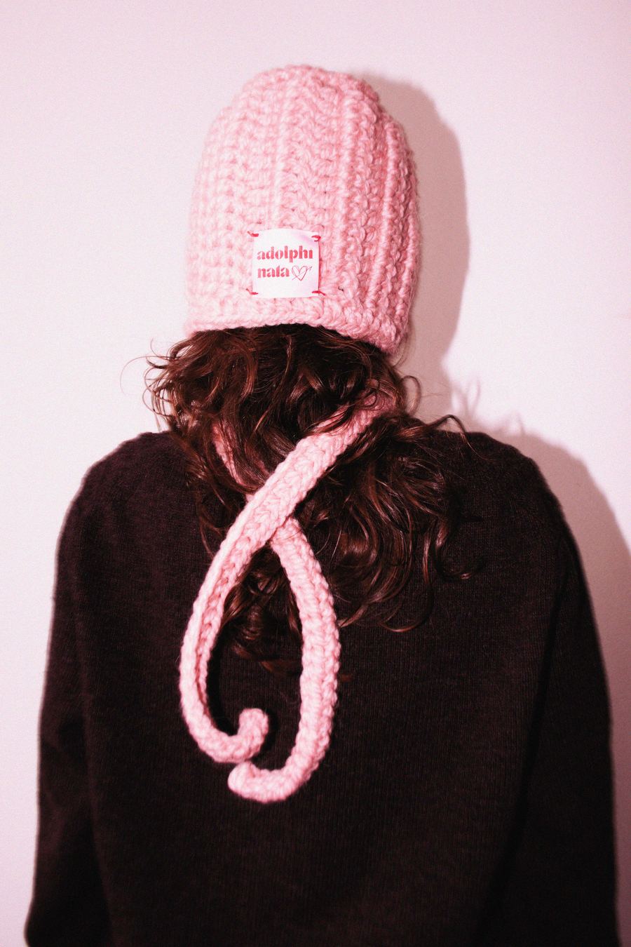 Rear view of a model in a pink ribbed knit beanie with an Adolphinata label, hair loose over a brown sweater.