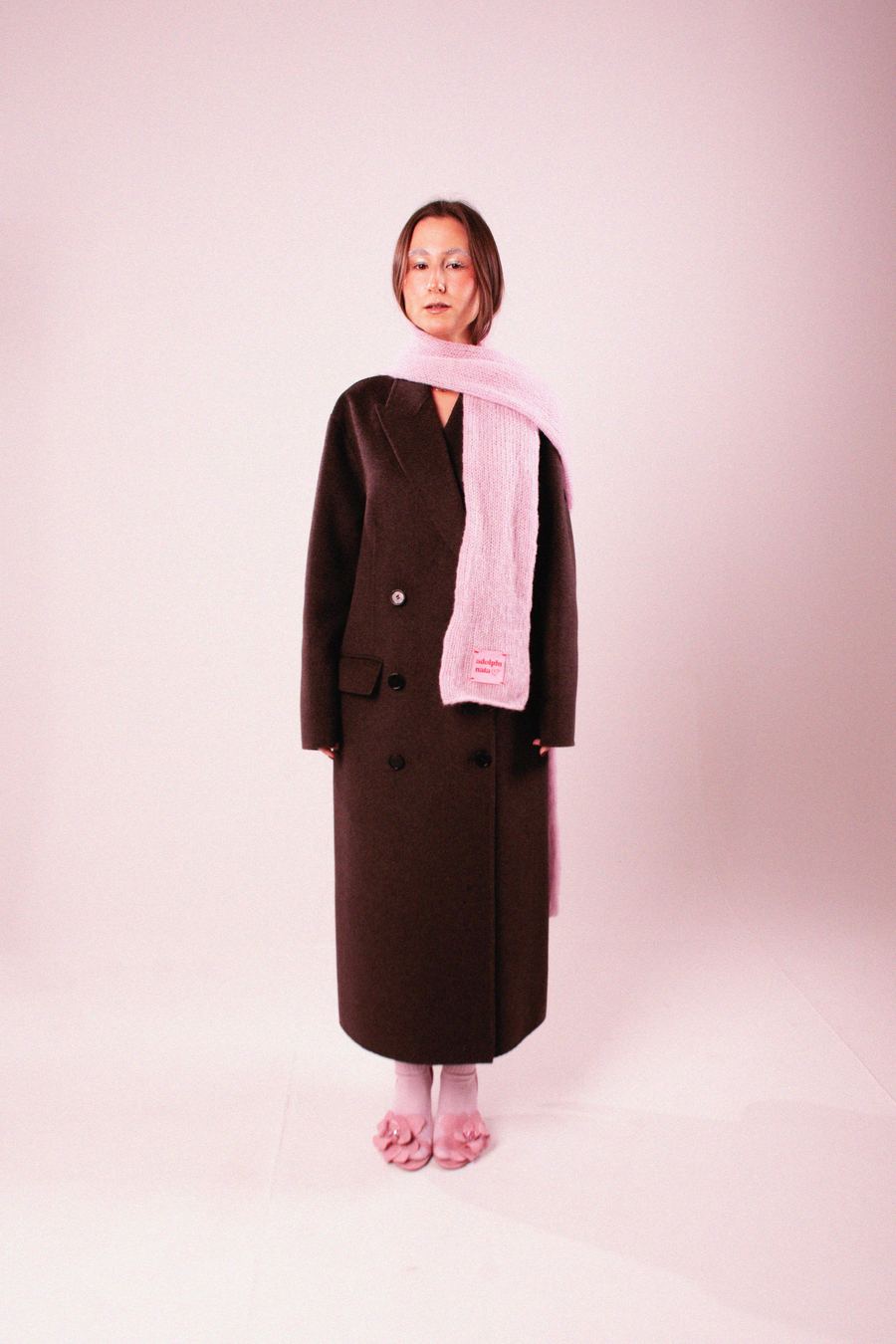 Model in a long brown double-breasted coat with a pink knit scarf draped over one shoulder, pink heeled sandals.